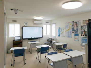 第1教室 壁に大型モニター、白い机に2つの椅子が1セットで4セット並んでいる