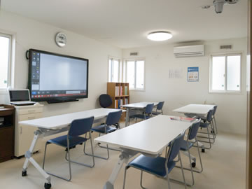 第2教室 壁に大型モニター、1人かけの白い机に椅子が1つずつ、5つ並んでいる